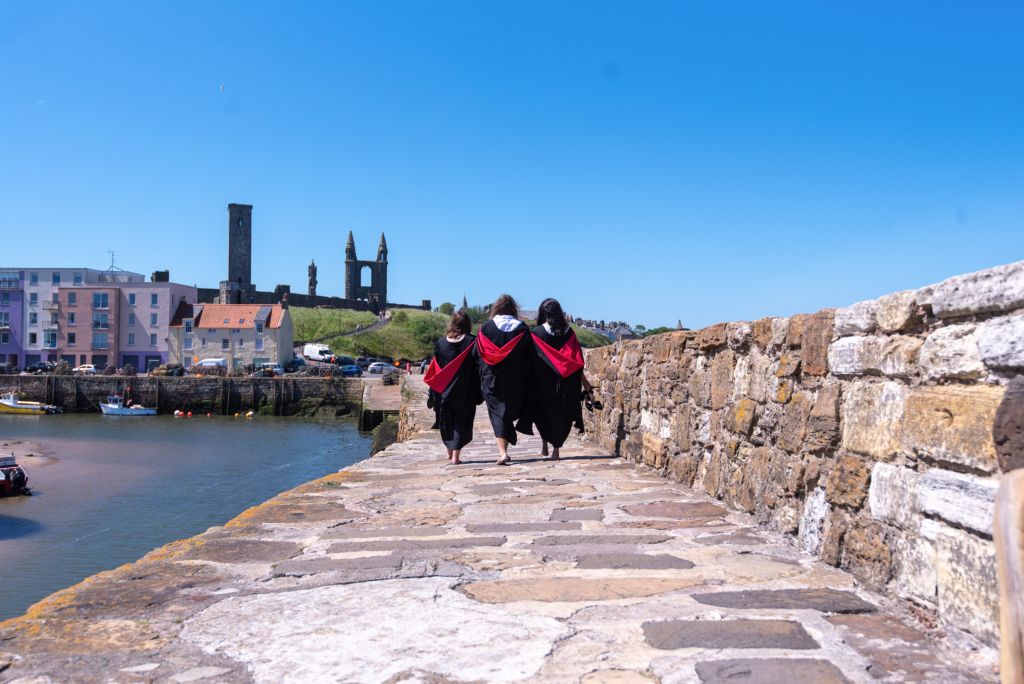 Three students wearing gowns and hoods walk on the St Andrews Pier.