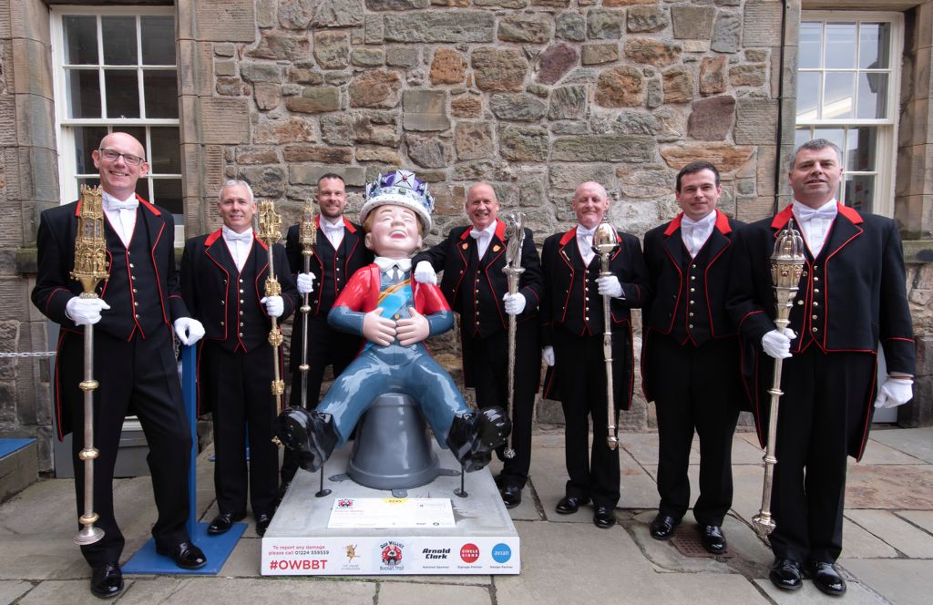 Seven mace bearers are standing in a line and smiling. They are holding the University Maces and posing beside an Oor Wullie statue.