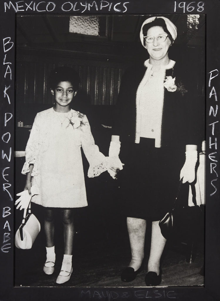 Black and white photograph shows a young black girl in white dress holding the hand of an older white lady in white shirt and black jacket. The pair are facing forwards.