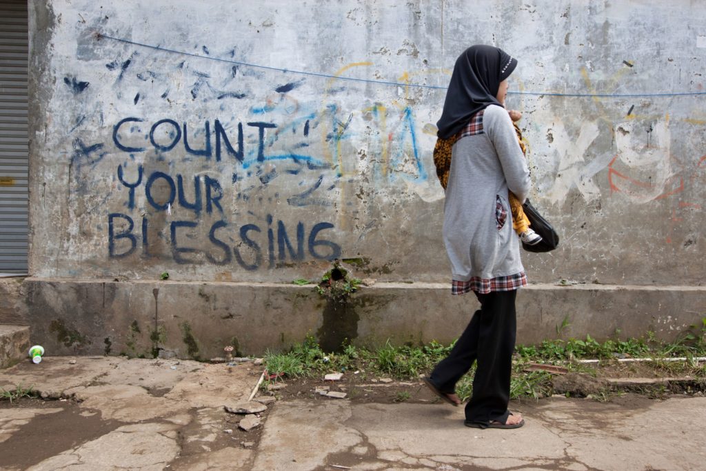 Colour photograph shows a woman holding a baby. The pair are facing to the right, walking away. They are in front of a wall which has 'Count your blessings' written on it in blue paint.