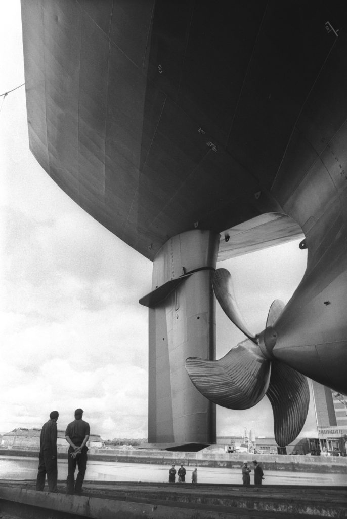 A black and white photo showing two men in a shipyard looking at the belly of a large ship. Other workmen are in the background.