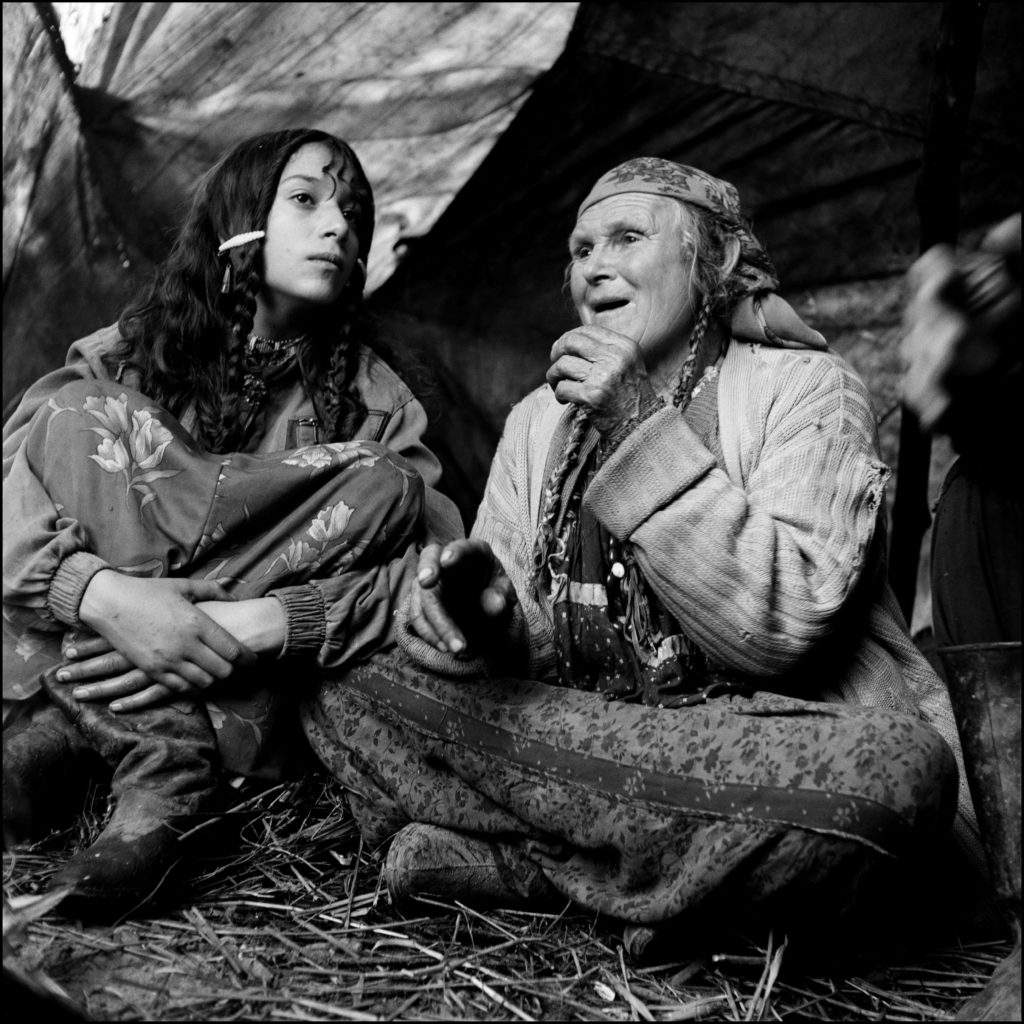 A black and white photo showing an elderly woman in traditional Romanian costume retells stories  to her granddaughter, sitting next to her on the left.