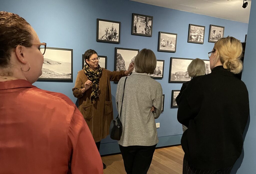 Some people are standing in a gallery listening to a tour, black and white photos hanging on the wall