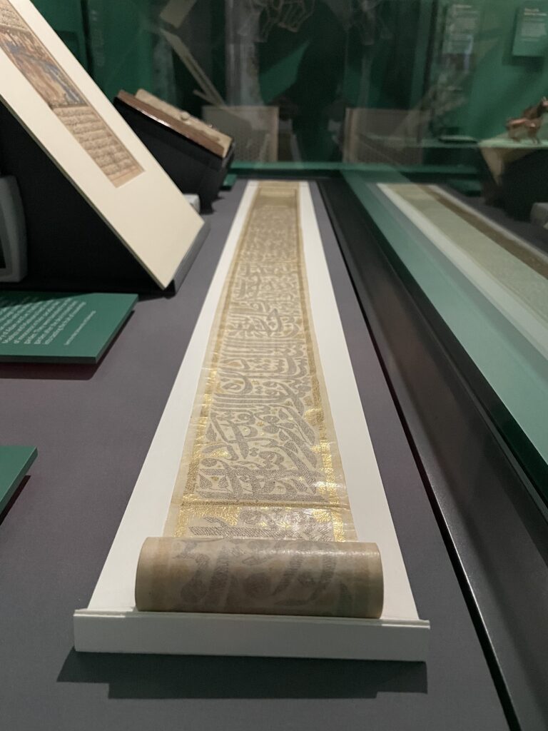 A small scroll unrolled on a white cardboard support. The miniature writing on the scroll is arranged to look like leaves and the edges are covered in gold.